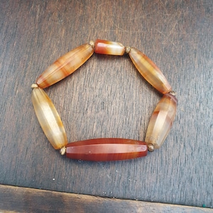 May include: A stretch bracelet composed of six elongated, faceted, translucent orange and brown agate beads. The beads are separated by small, round, metallic-toned spacer beads, creating a circular arrangement with a smooth, polished finish.