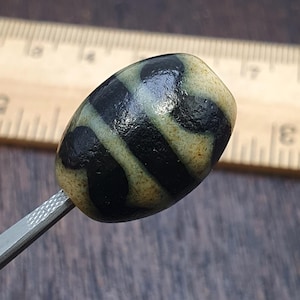 May include: A close-up of an oval-shaped bead with a cream-colored base and bold black stripes. The bead is held by a metal tool, with a wooden ruler in the background. The bead's design is symmetrical and eye-catching.