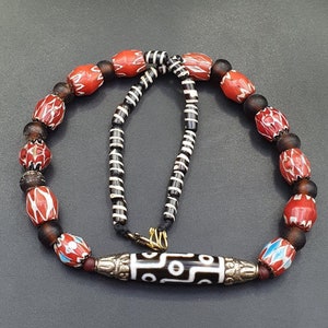 May include: A necklace made of red, black and white beads with a large, black and white agate pendant. The necklace is strung on a black cord and has a silver clasp.