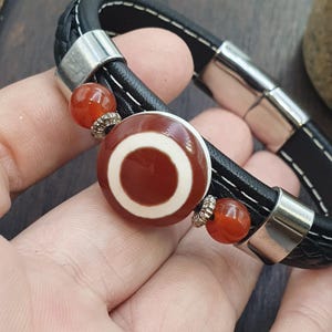 Puede incluir: Pulsera de cuero negro con una cuenta redonda central con un diseño marrón y blanco. La pulsera incluye dos cuentas naranjas y detalles metálicos plateados. La banda de cuero es trenzada y tiene costuras blancas.