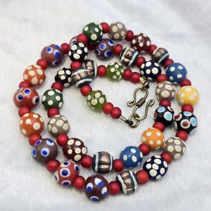 May include: A vibrant necklace with patterned glass beads in shades of orange, red, green, blue, and brown. The beads feature white polka dots and stripes. The necklace is strung on a red cord and has a brass clasp.