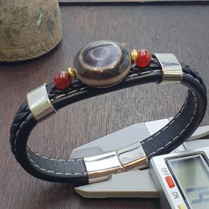 Puede incluir: Pulsera de cuero trenzado negro con una cuenta central estampada. La pulsera tiene detalles en rojo y dorado y un cierre plateado. Se muestra sobre una superficie de madera.