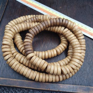 Puede incluir: Un largo collar de cuentas de madera circulares en forma de disco, en tonos beige claro y marrón oscuro. Las cuentas están ensartadas en un bucle continuo, y una regla visible en la parte superior muestra una longitud de 23 cm.