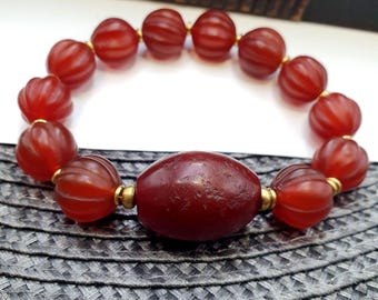 May include: A red beaded bracelet with a large oval bead in the center. The bracelet features smaller, round beads with a ribbed design. The beads are connected by gold-colored spacers. The bracelet is displayed on a textured, dark gray surface.