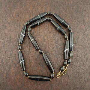 May include: A black beaded necklace with elongated beads and white stripes. The necklace has a gold-colored clasp and small gold-colored beads between the larger black beads. The necklace is displayed on a dark brown wooden surface.