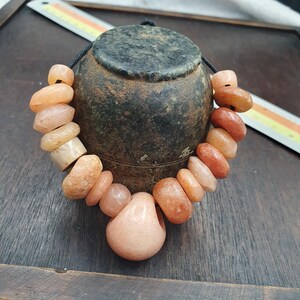 Puede incluir: Un collar con una serie de cuentas de piedra irregulares y lisas de color melocotón ensartadas en un cordón negro. Las cuentas varían en tamaño y forma, con una cuenta colgante más grande, redondeada y en forma de gota en el centro.