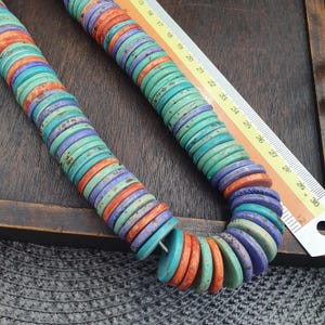 Puede incluir: Un collar de cuentas planas y circulares en turquesa, morado y naranja, con una textura moteada. Una regla está colocada junto a las cuentas, mostrando una medida de longitud que supera los 30 cm, indicando el tamaño del collar.