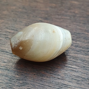 Könnte beinhalten: Ein glatter, oval geformter Stein, hellbeige mit braunen und weißen Bändern. Die polierte Oberfläche verleiht ihm ein natürliches Aussehen. Der Stein ist ein dekoratives Objekt. Etwa 5 cm lang.