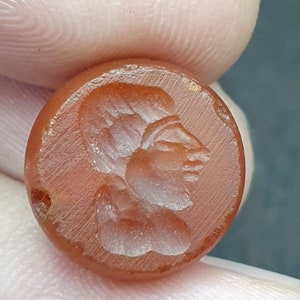 Pode incluir: Uma pedra redonda, castanho-avermelhada, esculpida com o perfil de um rosto humano. A escultura é detalhada, mostrando os traços faciais. Um pequeno orifício é perfurado na lateral, sugerindo que pode ser uma conta ou um pingente.