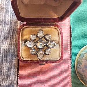 Op de afbeelding: Een zilveren ring met een cluster van heldere facetgeslepen stenen gezet in een stervormig ontwerp. De ring zit in een rode fluwelen ringdoos.