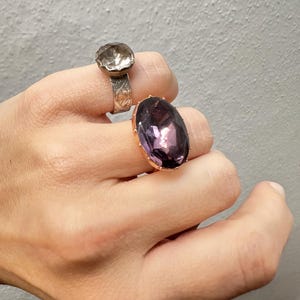 May include: Two rings are displayed on a hand. One ring has a silver band with a floral design and a clear, faceted stone. The other ring has a gold band and a large, oval-shaped, purple gemstone. The background is a plain, light gray wall.