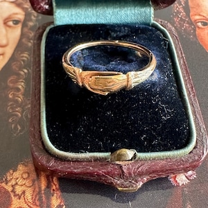 May include: A gold ring with an intricate design, presented in a dark blue velvet-lined jewellery box. The ring has a central rectangular section with leaf-like embellishments. The box is a deep burgundy colour.