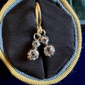 May include: A pair of antique gold earrings with two clear, round gemstones each. The earrings are displayed on a black velvet surface inside a blue and gold lined jewelry box. The earrings have a classic, elegant design.
