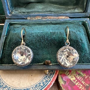Puede incluir: Un par de pendientes de plata con piedras de cristal facetadas transparentes. Los pendientes están en una caja de terciopelo verde.