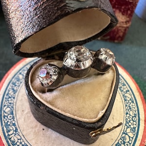 May include: A three-stone ring with a heart-shaped box. The ring has three large, clear stones set in silver.