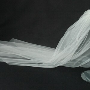 May include: A white tulle wedding veil displayed on a silver stand. The veil is long and flowing, with a delicate texture. A small tag with a butterfly design and the text "Silk Veil" is attached to the veil.