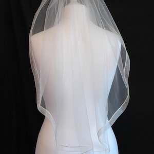 May include: A white bridal veil with a satin edge. The veil is on a white mannequin.