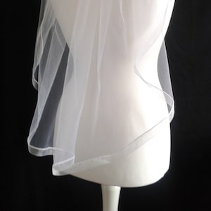 May include: A white single-tier bridal veil with a scalloped edge. The veil is made of soft tulle and is designed to be worn over the head.