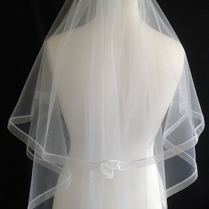 May include: A white, two-tiered bridal veil with a ribbon trim, displayed on a mannequin. The veil is sheer and delicate, with a simple bow detail at the waist. The background is black, highlighting the veil's details.