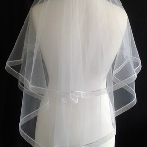 May include: A white bridal veil with a single layer of tulle and a satin trim. The veil is displayed on a white mannequin.