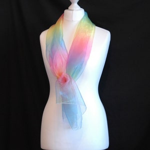 May include: A rainbow ombre sheer scarf draped on a white mannequin. The scarf is made of a lightweight, flowing fabric and features a gradient of colors from yellow to pink to blue.