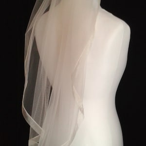 May include: A white bridal veil with a satin edge. The veil is draped over a white dress form.