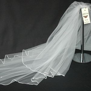 May include: A white bridal veil with a scalloped edge and small, sparkling embellishments. The veil is made of a sheer fabric and is displayed on a stand. A tag with the text "Little Vows" is attached.