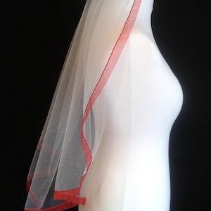 May include: White bridal veil with a red satin trim. The veil is on a white mannequin.