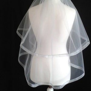 May include: A white single-tier bridal veil with a satin edge. The veil is displayed on a white mannequin.