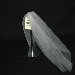 May include: A white tulle bridal veil displayed on a silver stand. The veil is made of soft, delicate fabric and has a flowing, elegant design. The tag attached to the veil reads "LB Veil, Designed for you by The Dressmaker."