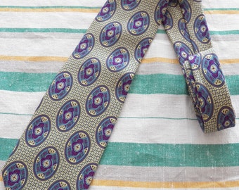 May include: A patterned necktie with a repeating oval design in shades of purple, blue, and yellow on a light beige background. The tie is made of a smooth fabric and is displayed on a striped fabric surface.