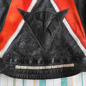 K&ouml;nnte beinhalten: Schwarze Leder-Motorradjacke mit roten und wei&szlig;en Streifen und einem schwarzen Dreiecks-Patch mit wei&szlig;em Logo. Die Jacke hat einen reflektierenden Streifen am unteren Rand.