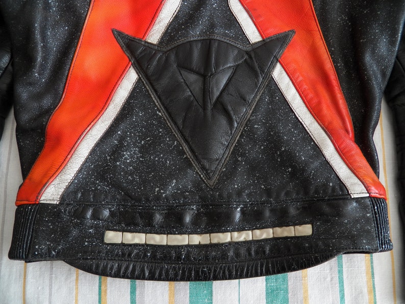 K&ouml;nnte beinhalten: Schwarze Leder-Motorradjacke mit roten und wei&szlig;en Streifen und einem schwarzen Dreiecks-Patch mit wei&szlig;em Logo. Die Jacke hat einen reflektierenden Streifen am unteren Rand.