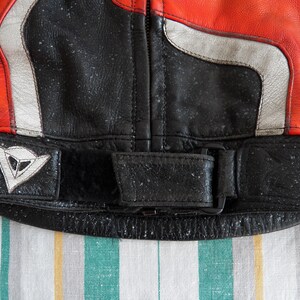K&ouml;nnte beinhalten: Nahaufnahme einer schwarzen Leder-Motorradjacke mit einem wei&szlig;en und roten Streifenmuster. Die Jacke hat ein Dainese-Logo auf der linken Seite und einen schwarzen G&uuml;rtel mit einer Schnalle.