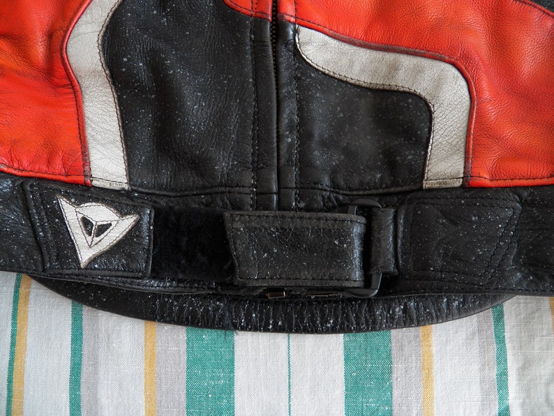 K&ouml;nnte beinhalten: Nahaufnahme einer schwarzen Leder-Motorradjacke mit einem wei&szlig;en und roten Streifenmuster. Die Jacke hat ein Dainese-Logo auf der linken Seite und einen schwarzen G&uuml;rtel mit einer Schnalle.