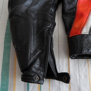 K&ouml;nnte beinhalten: Schwarze Lederjacke f&uuml;r Motorradfahrer mit roten und wei&szlig;en Akzenten. Die Jacke hat einen Rei&szlig;verschluss am linken &Auml;rmel und einen gerippten Bund am rechten &Auml;rmel.