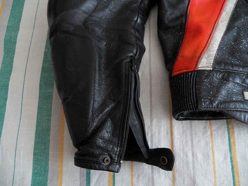 K&ouml;nnte beinhalten: Schwarze Lederjacke f&uuml;r Motorradfahrer mit roten und wei&szlig;en Akzenten. Die Jacke hat einen Rei&szlig;verschluss am linken &Auml;rmel und einen gerippten Bund am rechten &Auml;rmel.