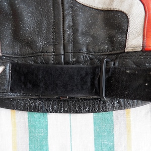 K&ouml;nnte beinhalten: Nahaufnahme einer schwarzen Lederjacke mit einem wei&szlig;en und roten Streifen-Detail. Die Jacke hat einen schwarzen verstellbaren Riemen mit einer Schnalle.