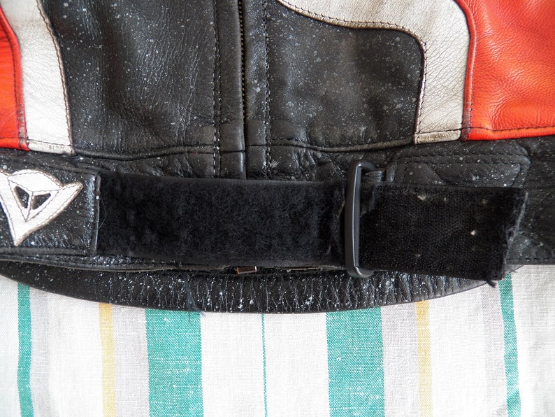 K&ouml;nnte beinhalten: Nahaufnahme einer schwarzen Lederjacke mit einem wei&szlig;en und roten Streifen-Detail. Die Jacke hat einen schwarzen verstellbaren Riemen mit einer Schnalle.
