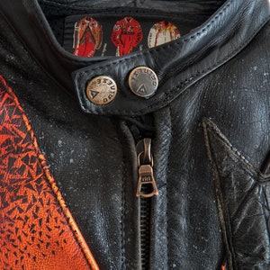 K&ouml;nnte beinhalten: Nahaufnahme einer schwarzen Leder-Motorradjacke mit orangefarbenen und roten geometrischen Mustern. Die Jacke hat einen Rei&szlig;verschluss und zwei Druckkn&ouml;pfe am Kragen. Das Etikett am Kragen lautet "Duesenberg".