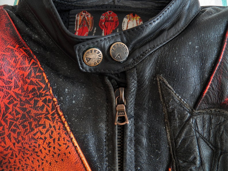 K&ouml;nnte beinhalten: Nahaufnahme einer schwarzen Leder-Motorradjacke mit orangefarbenen und roten geometrischen Mustern. Die Jacke hat einen Rei&szlig;verschluss und zwei Druckkn&ouml;pfe am Kragen. Das Etikett am Kragen lautet "Duesenberg".