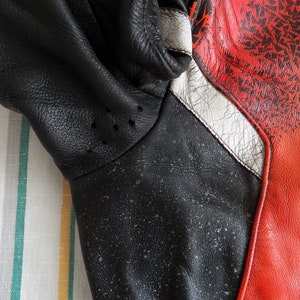 K&ouml;nnte beinhalten: Nahaufnahme einer schwarzen Lederjacke mit roten und wei&szlig;en Akzenten. Die Jacke hat eine strukturierte Oberfl&auml;che und ein Muster aus kleinen wei&szlig;en Punkten.