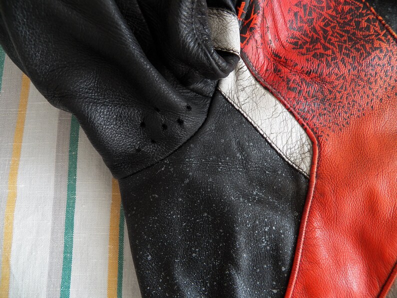 K&ouml;nnte beinhalten: Nahaufnahme einer schwarzen Lederjacke mit roten und wei&szlig;en Akzenten. Die Jacke hat eine strukturierte Oberfl&auml;che und ein Muster aus kleinen wei&szlig;en Punkten.