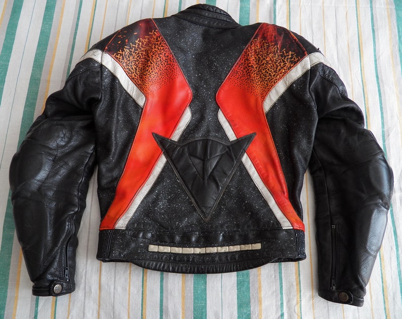 K&ouml;nnte beinhalten: Schwarze Leder-Motorradjacke mit roten, wei&szlig;en und orangefarbenen geometrischen Mustern. Die Jacke hat ein gro&szlig;es schwarzes Dreieck auf der R&uuml;ckseite mit einem wei&szlig;en Stern.