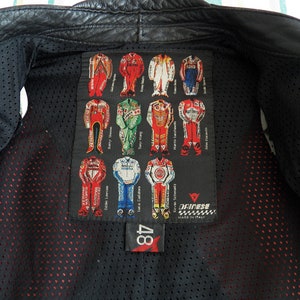 K&ouml;nnte beinhalten: Schwarze Leder-Motorradjacke mit einem roten und wei&szlig;en Dainese-Logopatch auf der R&uuml;ckseite. Der Patch zeigt ein Raster von Rennanz&uuml;gen mit den Namen von Fahrern wie Barry Sheene, Eddie Lawson und Luca Cadalora. Die Jacke hat ein Mesh-Futter und ein rotes Etikett mit der Nummer 48.