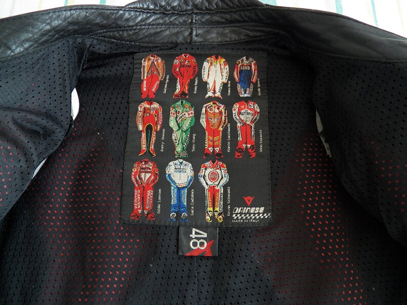 K&ouml;nnte beinhalten: Schwarze Leder-Motorradjacke mit einem roten und wei&szlig;en Dainese-Logopatch auf der R&uuml;ckseite. Der Patch zeigt ein Raster von Rennanz&uuml;gen mit den Namen von Fahrern wie Barry Sheene, Eddie Lawson und Luca Cadalora. Die Jacke hat ein Mesh-Futter und ein rotes Etikett mit der Nummer 48.