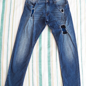 Może przedstawiać: Dżinsy z niebieskiego denimu z przetarciami i naszywką na prawej nogawce. Dżinsy mają prosty krój.