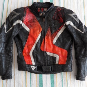 K&ouml;nnte beinhalten: Schwarze Leder-Motorradjacke mit roten und wei&szlig;en Akzenten. Die Jacke hat einen Rei&szlig;verschluss und ein Dainese-Logo auf der Vorderseite.