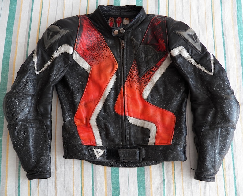 K&ouml;nnte beinhalten: Schwarze Leder-Motorradjacke mit roten und wei&szlig;en Akzenten. Die Jacke hat einen Rei&szlig;verschluss und ein Dainese-Logo auf der Vorderseite.