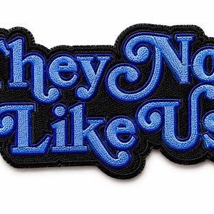 May include: A black embroidered patch with the words "They Not Like Us" in blue, stylized lettering. The patch has a black border and a textured appearance, suitable for application on clothing or accessories.